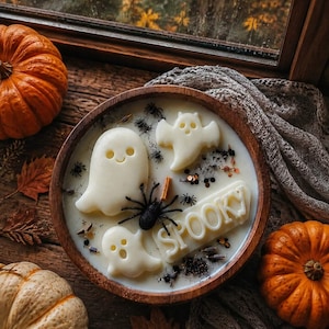 May include: A round wooden bowl filled with white wax featuring three white ghost-shaped wax embeds, a black spider figurine, and a white wax plaque with the word SPOOKY embossed on it. The wax is decorated with small dark botanical elements.