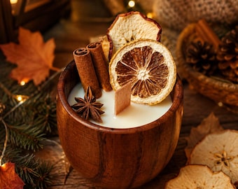 Könnte beinhalten: Eine weiße Wachskerze in einer dunkelbraunen Holzschale, verziert mit zwei Zimtstangen, einem Sternanis sowie getrockneten Orangen- und Apfelscheiben. Ein rechteckiger Holzdocht steht in der Mitte der glatten, cremeweißen Wachsoberfläche.