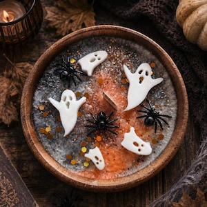 May include: A round wooden bowl filled with wax featuring a blend of gray and orange colors, embedded with white ghost-shaped wax pieces, black plastic spider figures, and gold hexagonal glitter, centered around a single wooden candle wick.