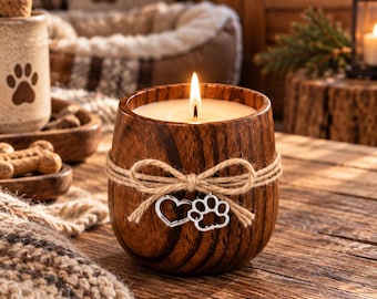 May include: A lit candle in a rounded dark wood container featuring a natural grain pattern. The container is wrapped with a tan jute twine bow, which holds two small silver-toned metal charms: one shaped like a heart and one like a paw print.