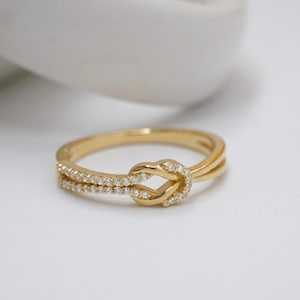 May include: A gold-colored ring with a knot design. The ring features a double band with small, clear stones set along one band. The knot is also adorned with the same small stones, creating a sparkling effect. The ring is set against a soft, white background.