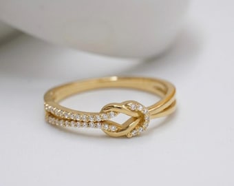 Puede incluir: Un anillo dorado con un diseño de nudo. El anillo presenta una doble banda, con pequeñas piedras transparentes engastadas a lo largo de una banda. El nudo también está adornado con las mismas pequeñas piedras, creando un efecto brillante. El anillo está sobre un fondo blanco suave.
