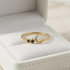 May include: A gold ring featuring a crescent moon shape and two small, round, black gemstones set side-by-side. The band is thin and polished, showcasing a smooth metallic finish. The ring is positioned inside a white, velvet-lined jewellery box.