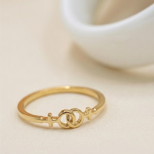 May include: A gold-colored ring with a double female symbol design. The ring is thin and delicate, with the symbols centered on the band. The ring is likely made of gold or a gold-colored metal.