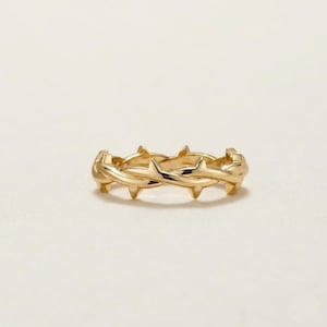 May include: A polished gold ring featuring a continuous band design that mimics the appearance of intertwined thorny vines. The metal surface is smooth and reflective, showcasing sharp, pointed protrusions along the entire circumference of the ring.