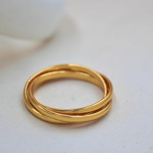 May include: A gold-colored ring with three intertwined bands. The ring has a smooth, polished surface and a bright, reflective finish. The bands are of equal width and create a seamless, interlocking design. The ring is set against a soft, neutral background.