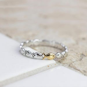 Könnte beinhalten: Ein silberner Ring mit einem sich wiederholenden Fischmuster, mit einem goldfarbenen Fisch. Der Ring ist vor einem neutralen Hintergrund platziert, der den Kontrast zwischen Silber und Gold hervorhebt. Ein zarter Schmuck.