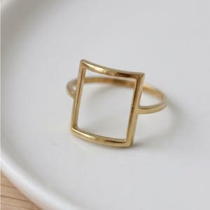 May include: A gold-colored ring with a square-shaped design. The ring features a thin band and a hollow square in the center. The ring is set against a white background.