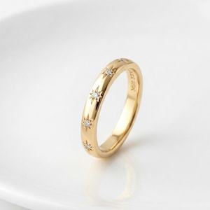 Könnte beinhalten: Ein goldener Bandring mit vier gleichmäßig verteilten sternförmigen Gravuren, die jeweils mit einem kleinen klaren Stein besetzt sind. Die Innenseite des Rings ist mit dem Text MSR 925 graviert. Der Ring hat eine polierte Oberfläche und ein abgerundetes Profil.