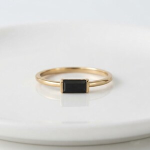 Puede incluir: Un anillo de oro con una piedra negra rectangular de corte baguette montada horizontalmente en una banda de metal sencilla. El anillo tiene un acabado pulido y un diseño limpio con la piedra oscura centrada en la banda fina y lisa.