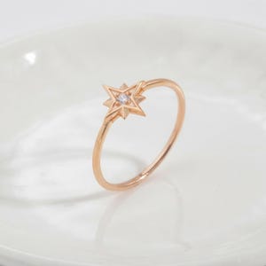 May include: A rose gold ring featuring a thin, polished band and a central star-shaped motif. The star design has sharp, pointed edges and is set with a single, small, clear round gemstone at its center, creating a delicate and refined appearance.