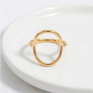 Puede incluir: Un anillo dorado con un diseño en forma de óvalo. El anillo está sobre una superficie blanca. La forma ovalada es el punto focal del diseño del anillo. Una joya sencilla y elegante.