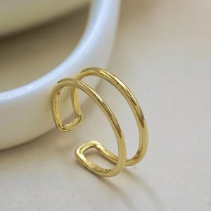 May include: A gold-coloured, open-ended ring with a double-band design. The ring has a smooth, polished surface and a minimalist style. The ring is likely made of metal and is shown against a neutral background.