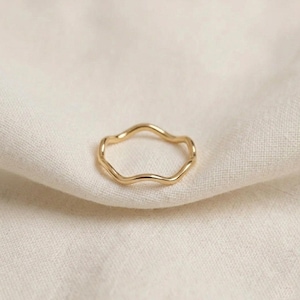Puede incluir: Un anillo dorado con un diseño ondulado y orgánico. El anillo se presenta sobre un fondo de tela de tono neutro, resaltando su forma delicada. La superficie lisa y pulida del anillo refleja la luz, creando un brillo sutil. Una pieza de joyería minimalista.