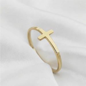 Peut inclure: Une bague dorée dotée d'un anneau fin et lisse, surmontée d'un petit emblème en forme de croix plate. Le métal présente une finition polie et réfléchissante, et le design est simple, composé d'un anneau circulaire avec une croix fixée.