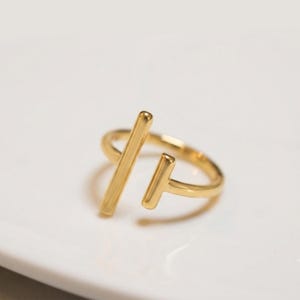Puede incluir: Un anillo abierto de tono dorado que presenta dos barras verticales paralelas de diferentes longitudes. El anillo tiene un acabado liso y pulido, con una banda circular que permanece abierta en la parte superior, creando un espacio entre los elementos.