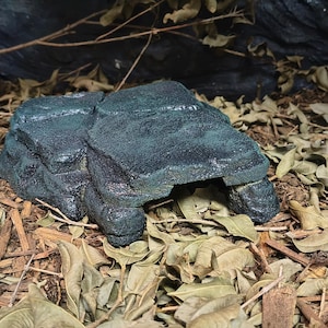 May include: A dark green, textured reptile hide cave shaped like a natural rock formation. The structure features a flat top surface with layered stone detailing and a wide, arched opening at the front, designed to provide a sheltered space.