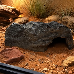 May include: A dark grey, textured rock-shaped hideout with a hollow opening on one side, positioned on a bed of reddish-brown sand. The structure features a rugged, uneven surface that mimics natural stone formations for a terrarium environment.