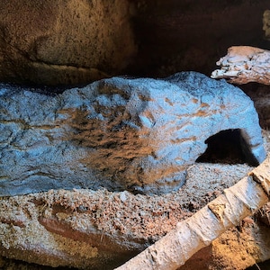 May include: A dark grey, textured reptile hide cave with a hollow opening, positioned on a sandy substrate next to light-coloured wooden branches. The structure features a rugged, rock-like surface with deep grooves and a realistic, uneven finish.