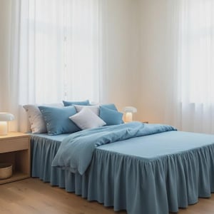 Puede incluir: Una cama con ropa de cama azul claro, que incluye una funda nórdica, almohadas y una falda de cama. La habitación tiene cortinas blancas transparentes y un suelo de madera. Dos pequeñas lámparas están en las mesitas de noche.