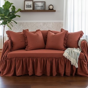 May include: A rust-colored sofa with matching throw pillows and a ruffled skirt. The sofa has a light blue and white fringed throw blanket draped over the arm. Decorative items are on a shelf in the background.