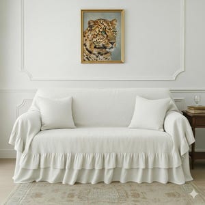 May include: A white sofa with a ruffled skirt and two matching square pillows. Above the sofa hangs a framed portrait of a leopard with green eyes. The room has a neutral color palette.