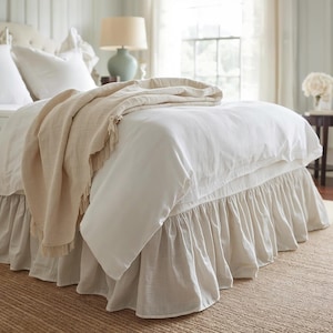 Könnte beinhalten: Ein Bett mit weißer Bettwäsche, einschließlich eines Bettbezugs und eines gerüschten Bettrands. Eine beige Decke mit Fransen ist über das Bett drapiert. Der Raum hat eine neutrale Farbpalette mit einer Lampe und einem Fenster im Hintergrund.
