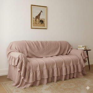 May include: A dusty rose sofa cover with ruffled edges. The cover drapes over a sofa, with a framed picture of a giraffe and elephant on the wall above. A small side table with books is visible.