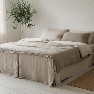 Może przedstawiać: Łóżko z pościelą lnianą w neutralnych odcieniach, w tym kołdrą, poduszkami i spódnicą łóżka. Łóżko znajduje się w jasnym pokoju z małym drzewkiem i drewnianym stolikiem nocnym. Na podłodze znajduje się tkany dywan.