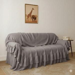May include: A gray sofa cover with ruffled edges. The cover is draped over a sofa, with a framed picture of a giraffe and elephant on the wall above. A small side table with books and a glass is next to the sofa.