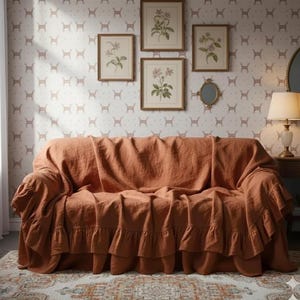 May include: A rust-colored sofa cover with a ruffled skirt draped over a sofa. The cover is made of a soft fabric and has a relaxed, casual look. The background features framed botanical prints and a patterned wallpaper.