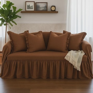 May include: A brown sofa with matching throw pillows and a ruffled skirt. The sofa has a cream-colored throw blanket draped over the arm. The room has a wooden shelf with framed pictures and a clock.