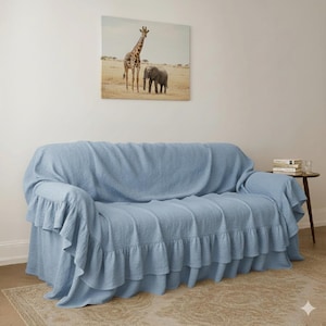 May include: A light blue sofa cover with ruffled edges. The cover is draped over a sofa, with a painting of a giraffe and elephant on the wall above. A small side table with books and a glass is to the right.