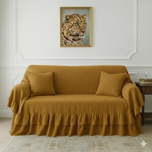 May include: Mustard yellow sofa with two matching throw pillows. The sofa is covered with a ruffled slipcover. A framed leopard portrait hangs above the sofa, adding a touch of wildlife to the room.