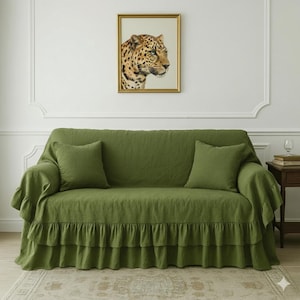 May include: A green sofa with two matching throw pillows and a ruffled skirt. Above the sofa hangs a framed portrait of a leopard. The sofa is covered with a green linen fabric.