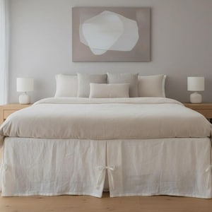 Może przedstawiać: Sypialnia z łóżkiem w neutralnych tonacjach. Łóżko ma beżową kołdrę i białą spódnicę. Na łóżku ułożono kilka poduszek w odcieniach beżu i bieli. Dwie białe lampy stoją na drewnianych stolikach nocnych. Nad nim wisi abstrakcyjne dzieło sztuki.