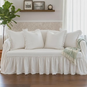 May include: A white sofa with ruffled skirt and matching throw pillows. A light green and white fringed throw blanket rests on the arm. Decorative items are on a shelf in the background.