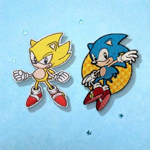 May include: Two embroidered patches featuring the character Sonic the Hedgehog. One patch shows a yellow Super Sonic in a dynamic pose with red and white shoes. The second patch depicts a blue Sonic leaping in front of a yellow checkered circle.
