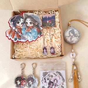 May include: A cardboard box filled with anime-style merchandise including a large acrylic standee of two characters, two small phone charms, a square print, two keychains, a plastic bag of stickers, and a circular hanging ornament with a gold tassel.