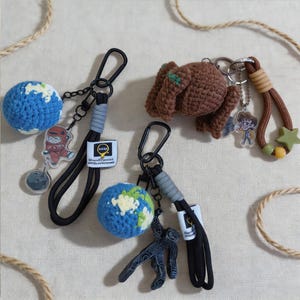 Peut inclure: Trois porte-clés composés de sphères crochetées représentant la Terre, d'une figurine crochetée marron et de breloques en acrylique incluant un astronaute, une personne et une silhouette noire. Chaque pièce a un mousqueton et un cordon noirs.