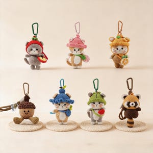 May include: A collection of seven crocheted keychains featuring various characters including cats wearing fruit-themed hats, a brown acorn with a smiling face, and a red panda, each equipped with a metal lobster clasp for attachment.
