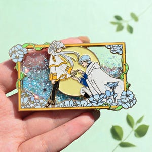 以下が含まれることがあります： 金色のフレームと青い花のアクセントが特徴の長方形のエナメルピン。中には長い白髪のキャラクターが立ち、もう一人が膝をついてその手を取っている様子が描かれています。背景は黄色い月と青いラメ入りの液体です。