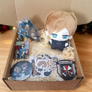 May include: A cardboard gift box filled with shredded paper containing a plush doll with light brown hair and blue eyes, an acrylic standee, a rectangular card, a circular pin, several small stickers, and a shield-shaped Umbrella Corporation patch.