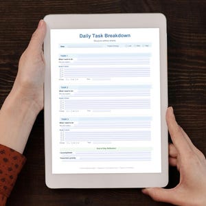 May include: A white tablet displaying a "Daily Task Breakdown" form is held by hands. A brown notebook with a green pencil, a keyboard, and a clipboard with sticky notes are on a dark wooden surface.