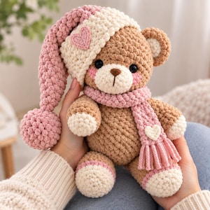 Könnte beinhalten: Ein gehäkelter Teddybär mit hellbraunem Fell, der eine cremefarbene und rosafarbene lange Schlafmütze mit Herzaufnäher sowie einen passenden rosafarbenen Schal mit einem kleinen weißen Herz trägt. Der Bär hat cremefarbene Pfoten und rosa Wangen.