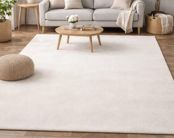 May include: A large, rectangular, solid off-white area rug with a soft, plush texture. The rug is positioned on a brown wood-plank floor in front of a light grey sofa, a wooden coffee table, and a woven pouf, creating a neutral floor covering.