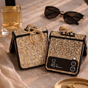 May include: Two bejeweled gold phone cases with black trim and gold keychains. The cases are covered in small, sparkling gold embellishments. A bottle of perfume and sunglasses are in the background.
