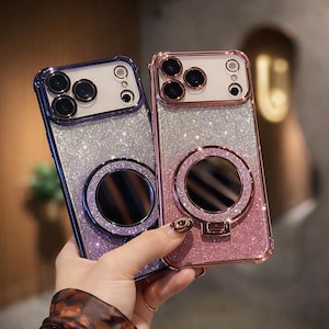 May include: Two glittery phone cases, one purple and one pink, are displayed. Each case has a circular mirror and a camera cutout. The cases have a sparkly, glittery design and a protective frame. The cases are held by a person.