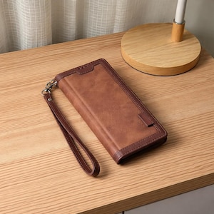 May include: A brown leather wallet with a wrist strap. The wallet has a rectangular shape with a darker brown border and a small tab closure. The wrist strap is also brown and attached with a silver clasp.
