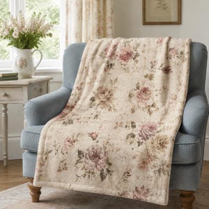 May include: A soft, cream-coloured throw blanket featuring a repeating pattern of muted pink and brown roses with green foliage. The blanket is draped over a blue upholstered armchair, showcasing a textured fabric finish and finished edges.